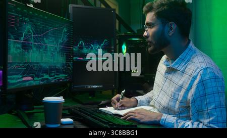 Indian Arabian man looking monitor computer chart graph trading online typing pc writing notes notebook analyst programmer male businessman business Stock Photo