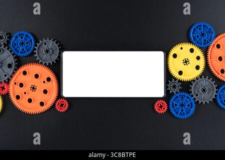 Top view of horizontal smartphone with blank white screen placed among colorful plastic gears on black surface. Ideal for concepts in technology, STEM Stock Photo