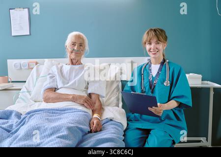 Senior Caucasian Woman Resting in Hospital Bed While Young Nurse Taking Notes Stock Photo