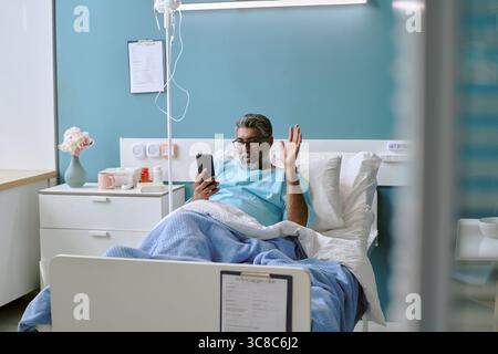 Middle Aged Biracial Man Lying in Hospital Bed Using Smartphone for Video Call Stock Photo
