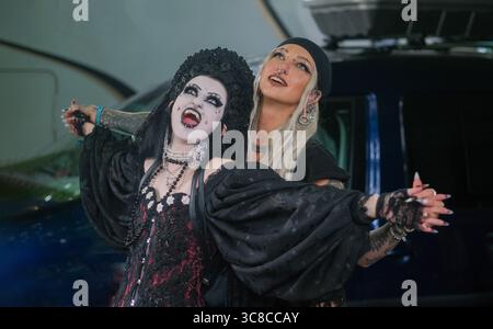 two gothic women pose playfully and expressively in elaborate makeup and dark clothing. one wears vampire fangs and a crown, the other has long blonde Stock Photo