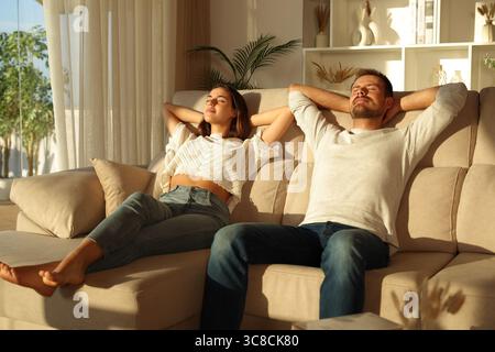 Happy couple relaxing with arms on head sitting on a couch at sunset at home Stock Photo