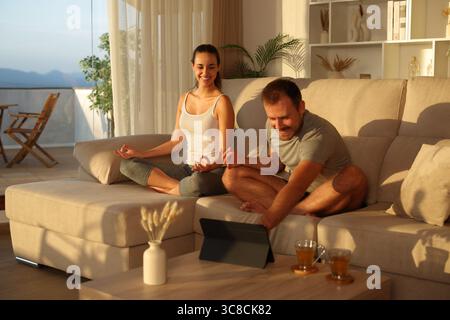 Happy couple preparing online yoga class on tablet sitting on a couch at sunset at home Stock Photo