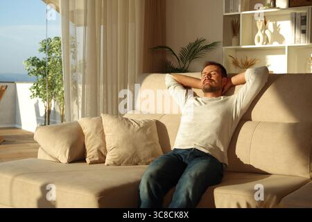 Man relaxing and resting with arms on head sitting on a sofa at home at sunset Stock Photo