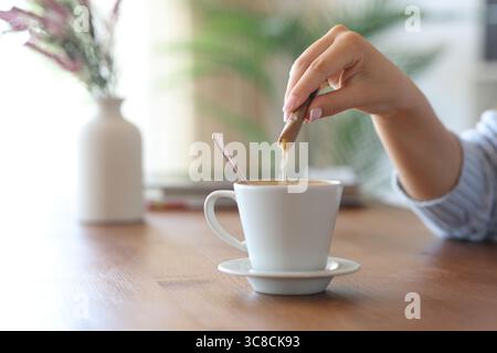 Close up of a woman hand throwing sugar from packet in a coffee cup on a wooden table at home Stock Photo