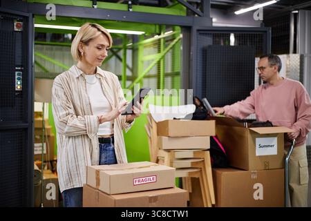 Caucasian Middle Aged Woman Using Digital Tablet While Packing Cardboard Boxes Stock Photo