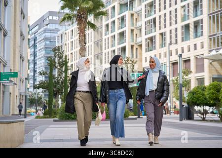 Three young muslim women wearing hijabs walking together in a modern city, talking and smiling Stock Photo