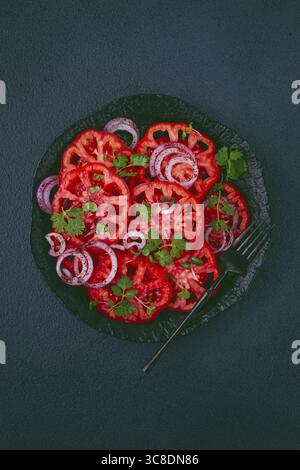 Freshly sliced tomatoes and red onions, garnished with herbs, on a black plate, summer salad, no people Stock Photo