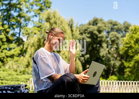 Urban stylish woman having online meeting using laptop while sitting on grass in park Stock Photo