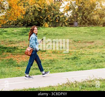 Full length portrait of young woman in denim attire walking outdoors in autumn city park. Stock Photo