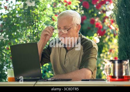 An elderly man sits with a laptop at a table in the garden. Stock Photo