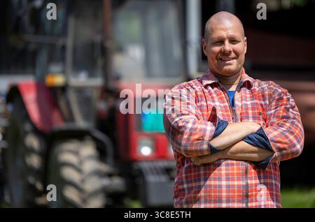 Male farmer is smiling confidently with arms crossed while standing in a field. Green tractor is visible in the background Stock Photo