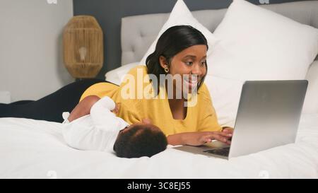 Smiling Mother Working on Laptop While Holding Her Baby on a Comfortable Bed Stock Photo