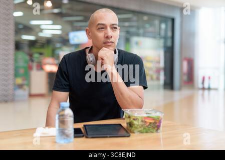 Mixed-race bald adult man in black t-shirt eating takeaway salad in mall food court. Healthy lifestyle, clean eating, and nutrition in real life setti Stock Photo