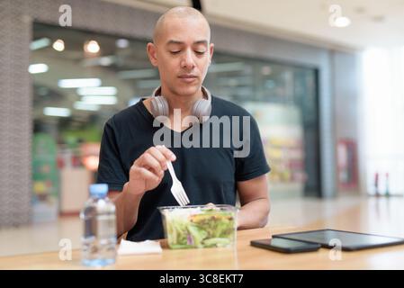 Mixed-race bald adult man in black t-shirt eating takeaway salad in mall food court. Healthy lifestyle, clean eating, and nutrition in real life setti Stock Photo