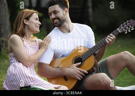Handsome man playing guitar for his pretty girfriend while they sit in a park, smiling and looking at each other Stock Photo