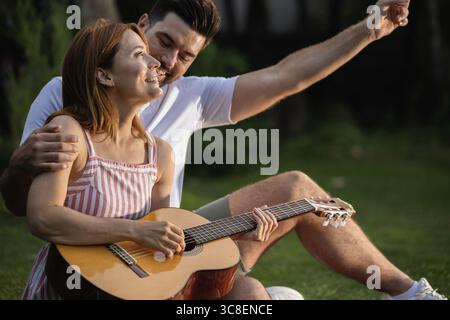Man pointing to a sunset while looking at his girlfriend that plays a guitar. The partners admire the sky while sitting in a park Stock Photo