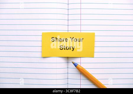 A yellow paper note with the words Share Your Story on it lying in an open notebook near a ballpoint pen. Close up. Stock Photo
