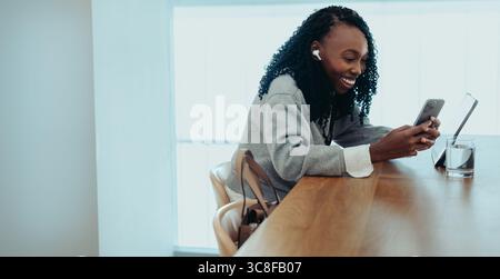 A smiling African businesswoman using her smartphone at a bright and modern workspace. She exudes relaxation and productivity, wearing wireless earbud Stock Photo