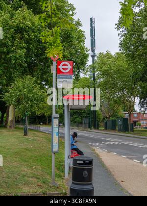 Man using mobile phone at bus stop with large 5G mobile communications mast visible in background, Chingford, London, UK Stock Photo