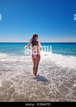Rear view beautiful woman in bikini stands in the foamy waves of the clear blue sea, facing the wide horizon under a cloudless summer sky. Summertime Stock Photo