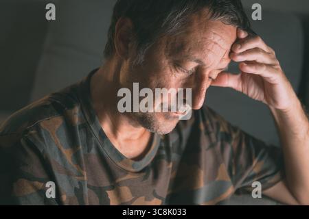 Stressed mature man with hand on forehead sitting indoors, dealing with anxiety or burnout, selective focus Stock Photo
