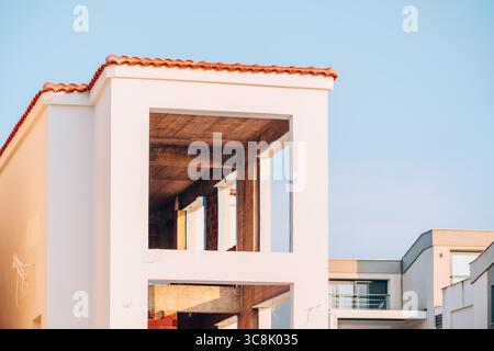 Minimalist view of an unfinished modern building with open concrete interior, white exterior walls, and terracotta roof tiles under clear blue sky. Se Stock Photo