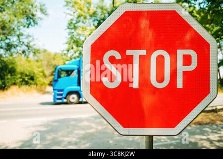 Bright red stop sign in sharp focus on a sunny day, with a blurred blue truck passing by in the background. Set against green trees and a quiet road. Stock Photo