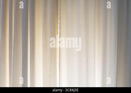 Sheer beige curtains with vertical stitching details softly illuminated by natural daylight. Minimalist interior decor with calming, warm tones.Abstra Stock Photo