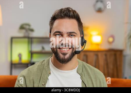 Confident young Caucasian man sitting on sofa at home wearing headset, freelance worker, call center, support service operator helpline, having talk with client or colleague, communication support. Stock Photo