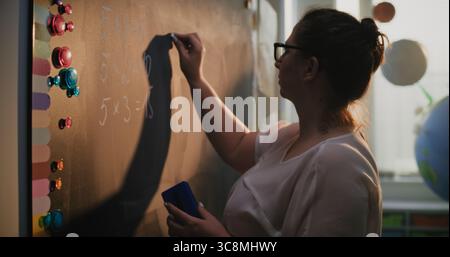 Female Teacher Writing Numbers and Multiplication Table on the Chalkboard, Teaching Primary School Students to Solve Math Problems During Lesson. Mathematical Class in Colorful Classroom. Close Up. Stock Photo