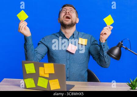 Exhausted man sitting at table with pasted sticker notes use laptop computer having concentration problem creative crisis. Nervous adult guy confused by big routine work loading on blue background Stock Photo