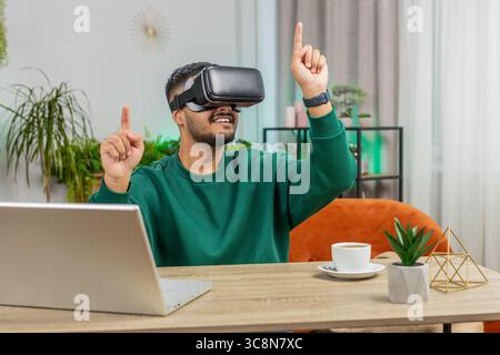 Indian man using virtual reality futuristic technology VR app headset helmet to play simulation 3D 360 online video game, watching film movie at modern home apartment. Guy in goggles sitting at desk Stock Photo