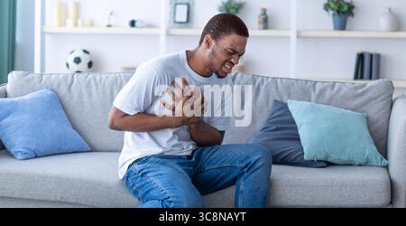 Millennial African American guy having heart attack, touching his chest in pain, sitting on sofa at home Stock Photo