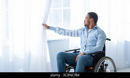 Sad with disability young man sitting on wheelchair alone at home, looking through window Stock Photo