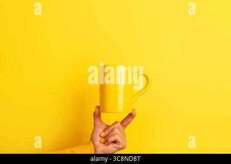 Hand in yellow sweater balancing a plain yellow ceramic mug on fingertips against a bright yellow background. Stock Photo