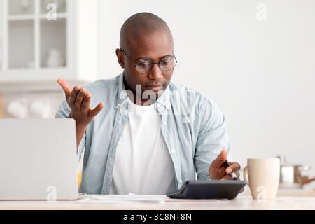 Savings and finances concept. Shocked black mature man calculating bills, using calculator, sitting in kitchen Stock Photo
