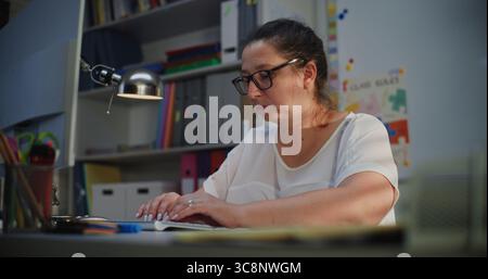 Elementary School Teacher Working on Computer, Checking Homework and Online Assignments During Distance Learning, Preparing Lesson Plan for Students. Woman Working at PC in Empty Classroom in Evening. Stock Photo