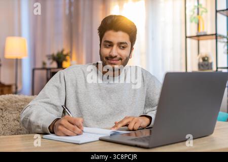 Young Indian man sitting at table joins online class through laptop, attentively listening to lecture, taking notes in notebook. Hispanic guy at home, focused on own task or learning activity nearby. Stock Photo