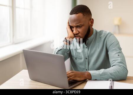 Unhappy Black Businessman Looking At Laptop Sitting At Workplace Indoor Stock Photo