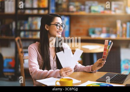Young creative graphic designer with color sets at cafe, using laptop and drinking coffee, empty space Stock Photo