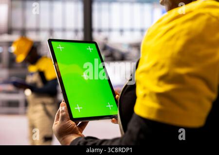 Factory manager handling operations as she holds tablet with green screen display. Professional industrial technician assessing production efficiency using digital device with mockup template. Stock Photo