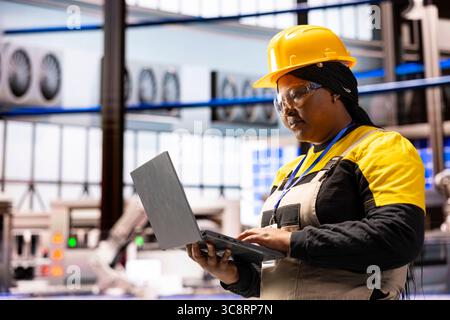 African american engineer with laptop working on manufacturing machines in industrial facility. Female factory supervisor using personal computer, overseeing production and doing quality control. Stock Photo