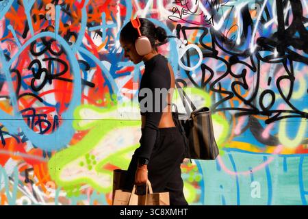 A young woman with headphones face partially in shade carrying shopping bags looking down on the ground against a busy graffiti background. Stock Photo