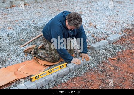 Worker carefully lays bricks for pathway, ensuring level placement on gravel surface during fall season. Stock Photo