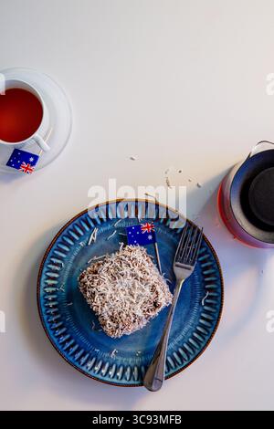 A Lamington cake with two Australian flags, one on the cake and one on a teacup saucer. A fork, blue plate, and glass teapot are on the white table. Stock Photo