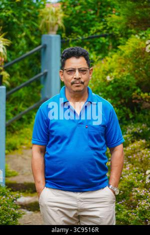 Man Standing in a Lush Green Garden with Colorful Flowers and Dense Foliage, Wearing Blue Shirt and Cream Pants. Stock Photo