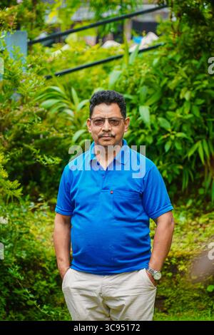 Man Standing in a Lush Green Garden with Colorful Flowers and Dense Foliage, Wearing Blue Shirt and Cream Pants. Stock Photo