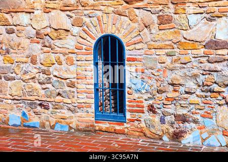 Ancient stonework reveals barred aperture. Small arched window with metal bars pierces a rugged stone wall, framed by bricks. Mix of stone sizes Stock Photo