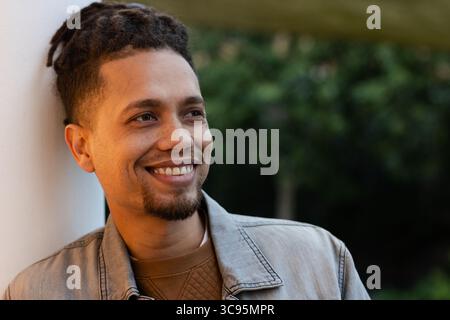 Mid adult African American man leaning against white column on outdoor patio, smiling gazing right Stock Photo